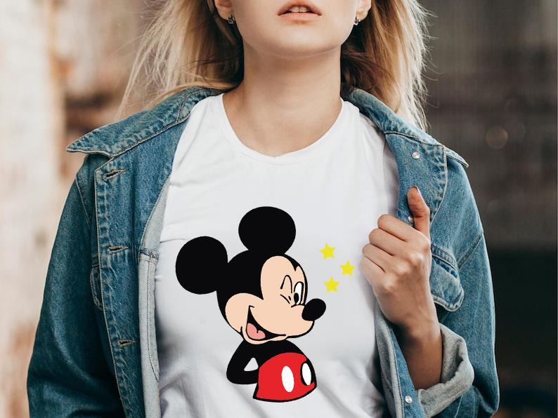 May include: White t-shirt featuring a smiling Mickey Mouse graphic with black ears, a red shorts, and yellow stars. The shirt is worn with a blue denim jacket.