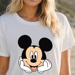 May include: White t-shirt featuring a smiling Mickey Mouse graphic. The cartoon character has black ears, a black face, and a tan face. Mickey's hands are white, and his tongue is pink. The shirt is a casual style.
