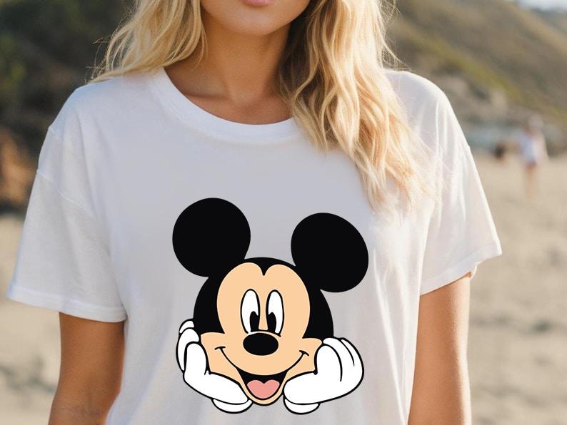 May include: White t-shirt featuring a smiling Mickey Mouse graphic. The cartoon character has black ears, a black face, and a tan face. Mickey's hands are white, and his tongue is pink. The shirt is a casual style.