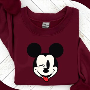May include: A maroon sweatshirt featuring a graphic of Mickey Mouse winking with his tongue sticking out. The design includes black ears and a white face with a red tongue. The sweatshirt has a crew neck and long sleeves.