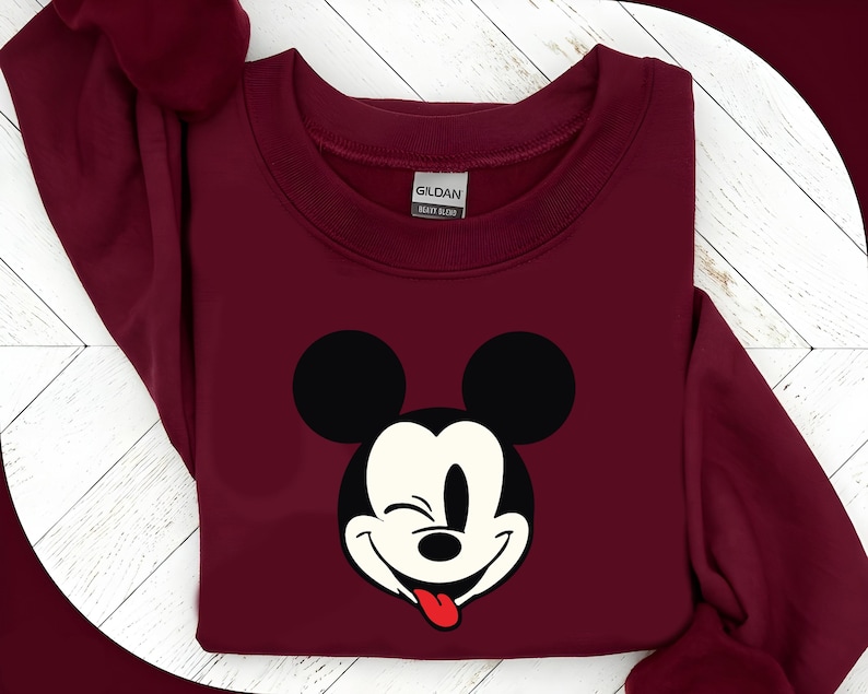 May include: A maroon sweatshirt featuring a graphic of Mickey Mouse winking with his tongue sticking out. The design includes black ears and a white face with a red tongue. The sweatshirt has a crew neck and long sleeves.