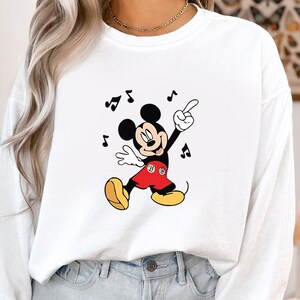 May include: White sweatshirt featuring a dancing Mickey Mouse graphic. Mickey is depicted in his classic attire: red shorts, yellow shoes, and white gloves. Black musical notes surround the character, suggesting a musical theme.