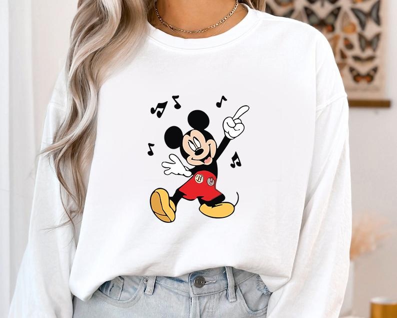 May include: White sweatshirt featuring a dancing Mickey Mouse graphic. Mickey is depicted in his classic attire: red shorts, yellow shoes, and white gloves. Black musical notes surround the character, suggesting a musical theme.