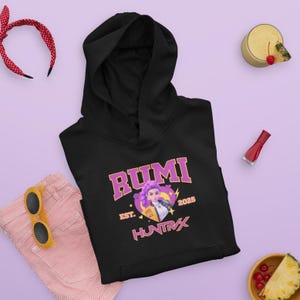 May include: Black hooded sweatshirt with the word "RUMI" in a pink and gold collegiate font. The sweatshirt features a graphic of a woman with purple hair and the text "EST. 2025 HUNTRX". The sweatshirt is displayed with a pair of pink striped pants and yellow sunglasses.