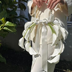 May include: White floral handbag with pearl accents and a satin bow. The bag features large, white, petal-like elements and green stems. The bag is held in front of a person wearing a white suit.