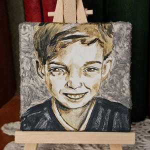 Könnte beinhalten: Ein kleines, quadratisches Gemälde einer lächelnden Person mit braunen Haaren und einem dunklen Hemd. Das Gemälde wird auf einer Miniatur-Holzstaffelei ausgestellt. Der Hintergrund ist strukturiert grau, und das Gemälde hat eine Vintage-Ästhetik.