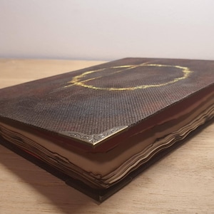May include: A leather-bound journal with a dark brown cover and gold-colored accents. The cover features a gold circle design. The corners are reinforced with decorative metal. The pages are aged, giving the journal a vintage look.