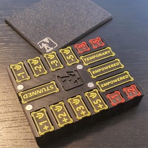 Könnte beinhalten: Ein schwarzes, 3D-gedrucktes Spiel-Tracker-Board mit einem zentralen Charakter-Symbol und magnetischen Schlitzen. Es enthält gelbe und rote rechteckige Einsätze mit Zahlen, Schwert-Symbolen und Texten wie STUNNED, TEMPORARY und EMPOWERED.