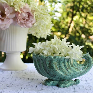 以下が含まれることがあります： 白い花が植えられた、貝殻の形をしたターコイズブルーのセラミック製のプランター。プランターは斑点模様の仕上げで、白いレースのテーブルクロスの上に置かれています。
