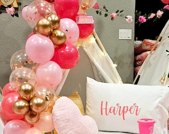 May include: A festive party scene with pink, gold, and clear balloons, a white pillow with "Harper" in pink script, and a pink heart-shaped pillow. A pink tumbler and sleep mask are also visible.