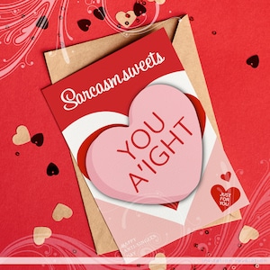 May include: A pink heart-shaped candy with the text "You A'ight" on it. The candy is on a red and white card with the text "Sarcasmsweets" and "Happy Anti-Singles Day" on it. The card is on a brown envelope with a red background.