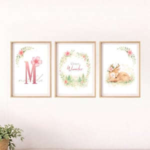 Op de afbeelding: Set van drie ingelijste kunstprints met een bloementhema. Eén print heeft de roze letter "M" en de naam "Matilda". Een andere print zegt "Kleines Wunder". De derde print toont een slapend hert met een bloemenkrans.
