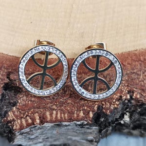 Puede incluir: Un par de pendientes de botón dorados. Cada pendiente presenta un diseño circular con un gráfico de baloncesto en el centro, rodeado por un anillo de pequeños cristales transparentes. Los pendientes se exhiben sobre una superficie de madera marrón texturizada.