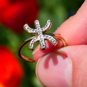 Op de afbeelding: Een goudkleurige ring met een gestileerd, opengewerkt ontwerp versierd met kleine, heldere edelstenen. De ring wordt vastgehouden tegen een vage achtergrond van rode en blauwe bloemen.