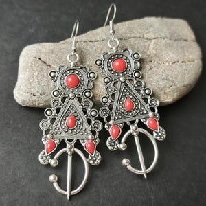 Puede incluir: Pendientes colgantes plateados con detalles rojos. Los pendientes presentan intrincados diseños geométricos con elementos circulares y triangulares, y una forma curva en forma de gancho en la parte inferior. Se exhiben sobre una piedra gris.