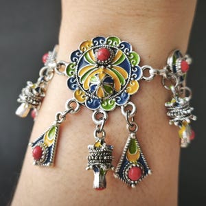 May include: A silver-toned charm bracelet featuring a central circular medallion with blue, yellow, and green enamel detailing and a small red bead. The bracelet includes dangling triangular and cylindrical charms, each accented with red beads and enamel.