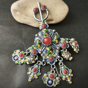 May include: A silver-toned brooch with a central red stone and intricate enamel detailing in blue, green, and yellow. The brooch features dangling pendants with red accents and is set against a dark background.