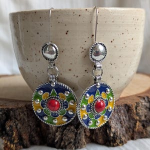 Puede incluir: Un par de pendientes colgantes plateados. Cada pendiente presenta un colgante circular esmaltado con fondo azul, detalles verdes, amarillos y blancos. Una piedra roja está engastada en el centro. Los pendientes se exhiben sobre una superficie de madera.