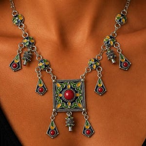 May include: A silver-toned necklace featuring a central square pendant with a red circular stone, surrounded by green and yellow enamel detailing. The chain includes multiple dangling charms with red stones and green and yellow enamel accents.
