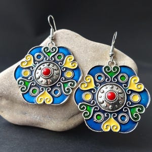 May include: A pair of ornate, floral-shaped earrings. The earrings feature a silver-colored metal base with a blue enamel background. The design includes yellow and green accents, and a red center stone. The earrings are displayed on a stone.