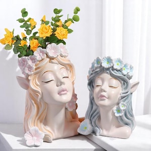以下が含まれることがあります： 2つの装飾的な頭の形をしたプランター。1つはブロンドの髪で黄色いバラが詰まっています。もう1つは灰色の髪で青い花の冠があります。どちらも目を閉じており、花のアクセントで飾られています。
