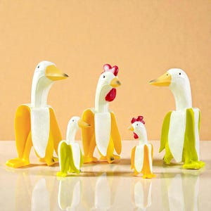Könnte beinhalten: Eine Sammlung skurriler Vogel-Figuren in Bananenform. Die Figuren zeigen Enten und Hühner mit gelben Bananenschalen und weißen und gelben Vogelkörpern. Die Vögel haben gelbe Schnäbel und rote Kämme. Die Figuren stehen auf einer reflektierenden Oberfläche.