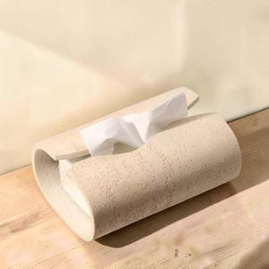 Puede incluir: Una funda para caja de pañuelos de color beige con una superficie texturizada y porosa. El soporte rectangular presenta un diseño curvo y abierto que envuelve una caja de cartón blanca, con un pañuelo de papel blanco saliendo por la ranura.