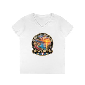 May include: A white V-neck t-shirt featuring a circular graphic on the chest. The design depicts a sunset over the French Riviera coastline with palm trees, boats, and the text Heritage Solaire Travel Club, 2026 and French Riviera.