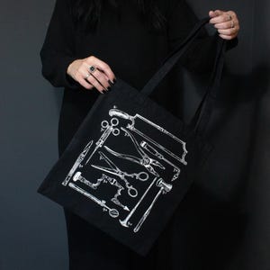 Schwarze Tragetasche mit einer weißen Illustration verschiedener chirurgischer Instrumente. Die Werkzeuge umfassen Sägen, Scheren und andere medizinische Instrumente. Die Tasche hat schwarze Griffe und besteht aus robustem Material.