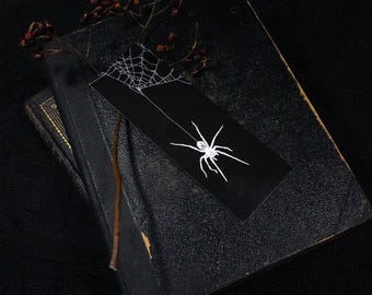 May include: A rectangular black bookmark featuring a white line art illustration of a spider hanging from a web. The bookmark is placed on top of a dark, textured, vintage-style book, accompanied by a thin brown twig with small dark red berries.