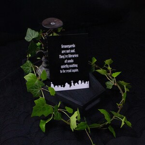 Eine schwarze Karte mit dem Text "Graveyards are not sad. They're libraries of souls quietly waiting to be read again." wird auf einer dunklen Holzkiste angezeigt, umgeben von grünem Efeu. Im Hintergrund ist ein dekoratives Metallobjekt zu sehen.