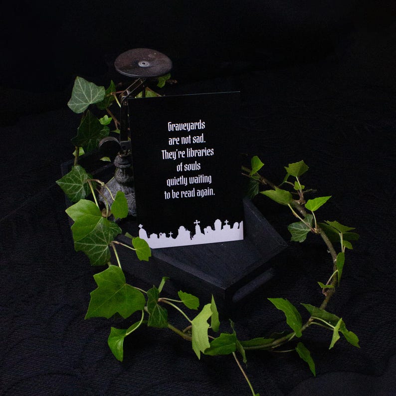 Eine schwarze Karte mit dem Text "Graveyards are not sad. They're libraries of souls quietly waiting to be read again." wird auf einer dunklen Holzkiste angezeigt, umgeben von grünem Efeu. Im Hintergrund ist ein dekoratives Metallobjekt zu sehen.
