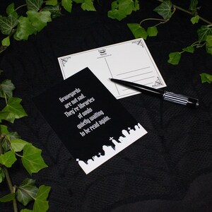 Eine schwarze Postkarte mit weißem Text: "Friedhöfe sind nicht traurig. Sie sind Bibliotheken der Seelen, die leise darauf warten, wieder gelesen zu werden." Weiterhin ist die Rückseite der Postkarte sichtbar.