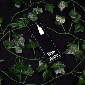 May include: A black bookmark with a white silhouette and the words "Night Reader." is surrounded by green ivy vines and leaves. The bookmark has a white border and a white silhouette of a figure.