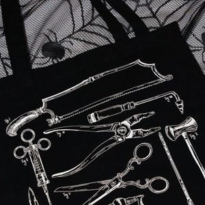Schwarze Tragetasche mit einer weißen Illustration antiker chirurgischer Instrumente, darunter Sägen, Zangen und Scheren. Im Hintergrund ist ein schwarzes Spinnennetz-Muster zu sehen.