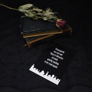 以下が含まれることがあります： 白い文字で「Graveyards are not sad. They're libraries of souls quietly waiting to be read again.」と書かれた黒い長方形のカード。テキストの下には様々な墓石の白いシルエットがある。近くには2冊の古い本と枯れたバラが置かれている。