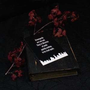 以下が含まれることがあります： 「Graveyards are not sad. They're libraries of souls quietly waiting to be read again.」という白い文字が書かれた黒い長方形のカード。テキストの下には白い墓地のシルエットがある。カードは使い古された黒い本の上に置かれている。