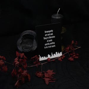 以下が含まれることがあります： 「Graveyards are not sad. They're libraries of souls quietly waiting to be read again.」という白い文字が書かれた黒い長方形のカード。カードの下部には、墓石や十字架が並ぶ墓地の白いシルエットが描かれています。