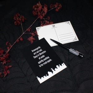 以下が含まれることがあります： Graveyards are not sad. They're libraries of souls quietly waiting to be read againという白い文字が書かれた黒いポストカード。下部には白い墓石のシルエットがあり、白黒のペンと無地のポストカードが添えられている。