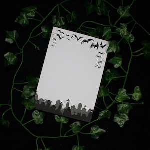 Könnte beinhalten: Eine weiße Karte mit Halloween-Motiv. Der obere Rand zeigt eine Silhouette von Fledermäusen im Flug, während der untere Rand eine Friedhofsszene zeigt. Die Karte ist von grünem Efeu vor einem schwarzen Hintergrund umgeben.