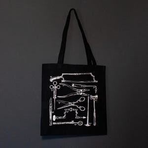 Schwarze Tragetasche mit einer weißen Illustration von chirurgischen Instrumenten im Vintage-Stil. Das Design umfasst Sägen, Scheren, Zangen und andere medizinische Werkzeuge. Die Tasche hat ein schlichtes, minimalistisches Design.
