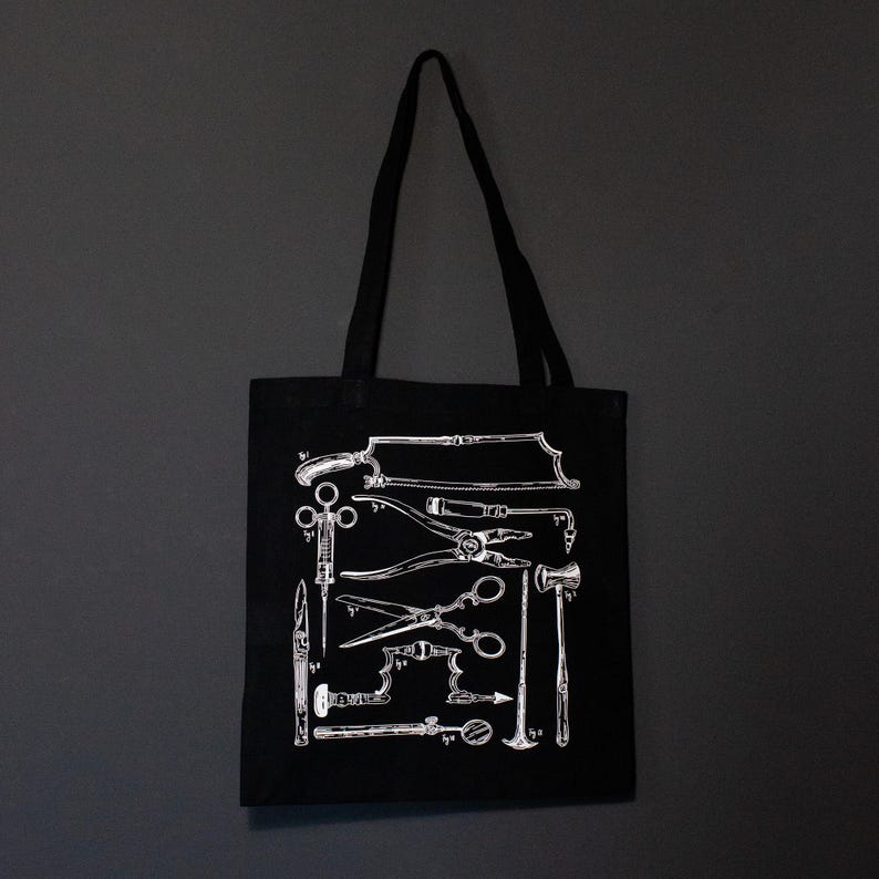 Schwarze Tragetasche mit einer weißen Illustration von chirurgischen Instrumenten im Vintage-Stil. Das Design umfasst Sägen, Scheren, Zangen und andere medizinische Werkzeuge. Die Tasche hat ein schlichtes, minimalistisches Design.
