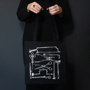Schwarze Tragetasche mit einer weißen Illustration von Vintage-Medizinwerkzeugen. Das Design umfasst Sägen, Scheren und andere chirurgische Instrumente. Die Tasche wird vor einem dunklen Hintergrund gehalten.