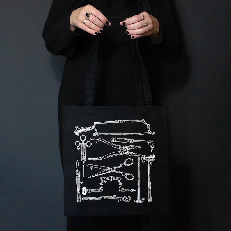 Schwarze Tragetasche mit einer weißen Illustration von Vintage-Medizinwerkzeugen. Das Design umfasst Sägen, Scheren und andere chirurgische Instrumente. Die Tasche wird vor einem dunklen Hintergrund gehalten.