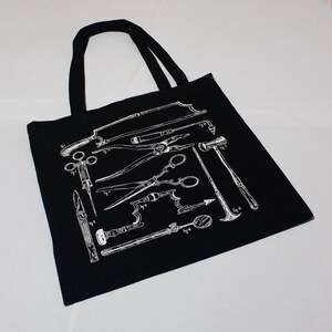 Schwarze Tragetasche mit einer weißen Illustration von chirurgischen Instrumenten im Vintage-Stil. Das Design umfasst Sägen, Scheren und Zangen. Die Tasche hat zwei schwarze Tragegriffe.
