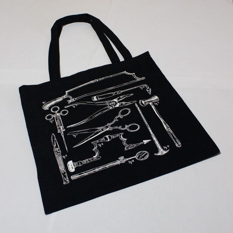 Schwarze Tragetasche mit einer weißen Illustration von chirurgischen Instrumenten im Vintage-Stil. Das Design umfasst Sägen, Scheren und Zangen. Die Tasche hat zwei schwarze Tragegriffe.