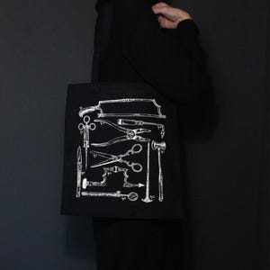 Schwarze Tragetasche mit einer weißen Illustration verschiedener chirurgischer Instrumente. Die Werkzeuge umfassen Sägen, Scheren und andere medizinische Instrumente. Die Tasche hat schwarze Griffe und besteht aus robustem Material.