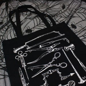 Schwarze Tragetasche mit einer weißen Grafik antiker chirurgischer Instrumente. Das Design umfasst eine Säge, eine Zange, eine Schere und einen Hammer. Die Tasche hat zwei schwarze Griffe und wird auf einem schwarz-weißen Spinnennetzstoff präsentiert.