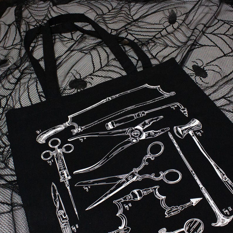 Schwarze Tragetasche mit einer weißen Grafik antiker chirurgischer Instrumente. Das Design umfasst eine Säge, eine Zange, eine Schere und einen Hammer. Die Tasche hat zwei schwarze Griffe und wird auf einem schwarz-weißen Spinnennetzstoff präsentiert.