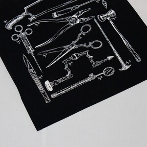 Schwarzer Stoff mit einer weißen Illustration von chirurgischen Werkzeugen im Vintage-Stil, darunter Sägen, Scheren und Zangen. Das Design zeigt detaillierte Strichzeichnungen verschiedener medizinischer Instrumente und schafft ein klassisches, anatomisches Thema.